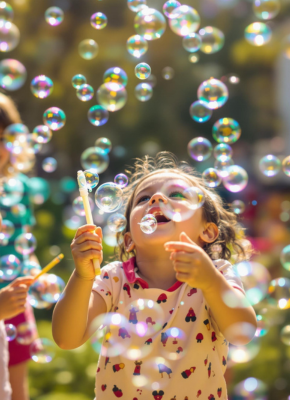 A young girl joyfully playing with soap bubbles outdoors, surrounded by colourful reflections in the sunlight.